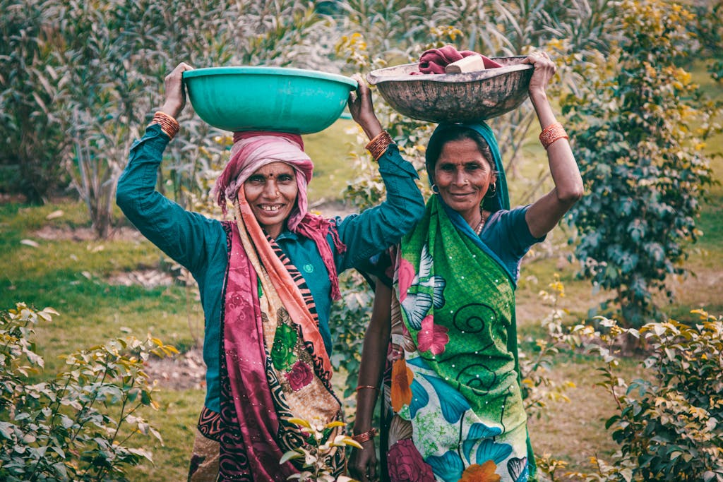 Two women wearing colorful traditional dresses smile outdoors, carrying baskets on their heads.