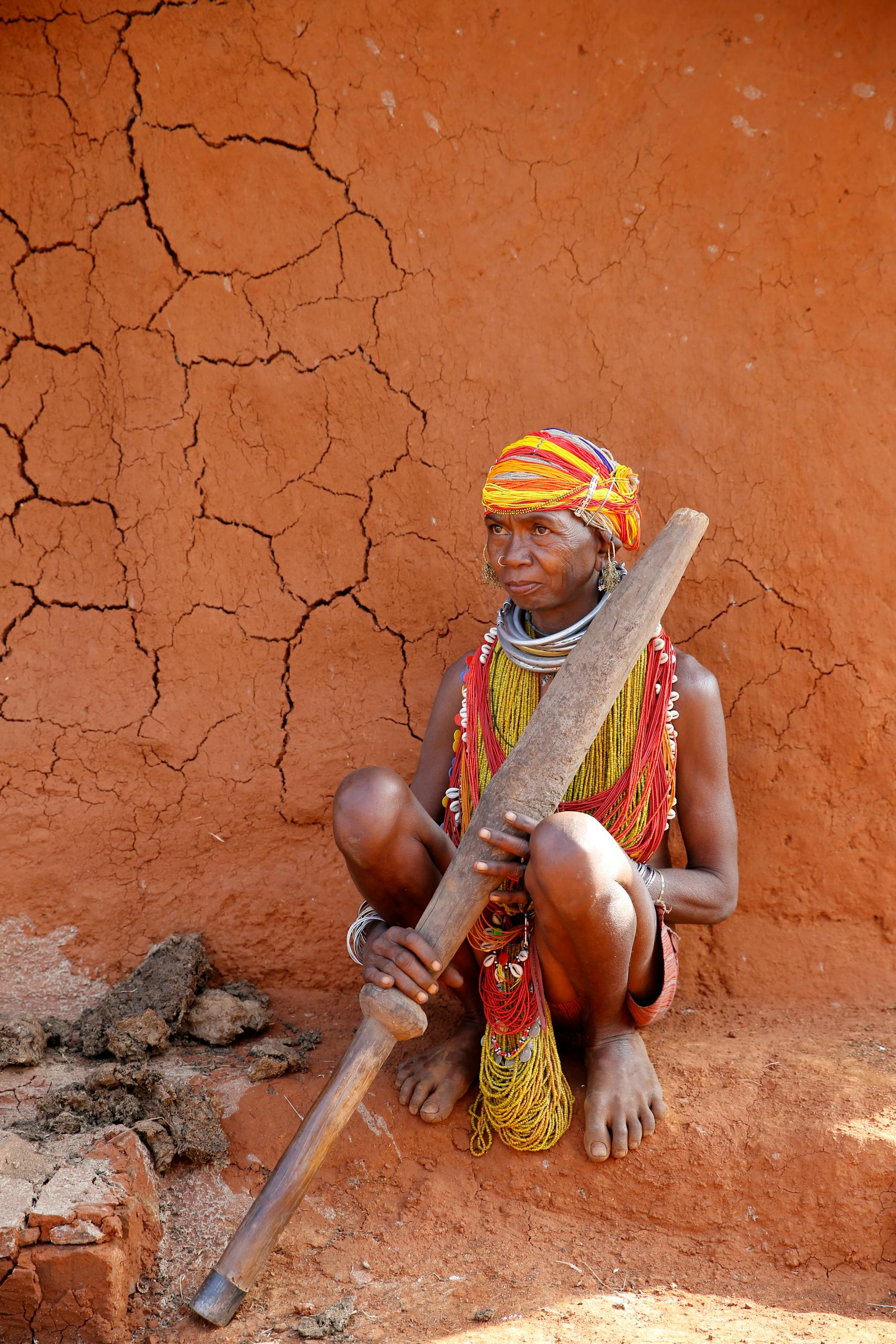A tribal woman in colorful traditional attire sitting by a mud wall in Odisha, India.